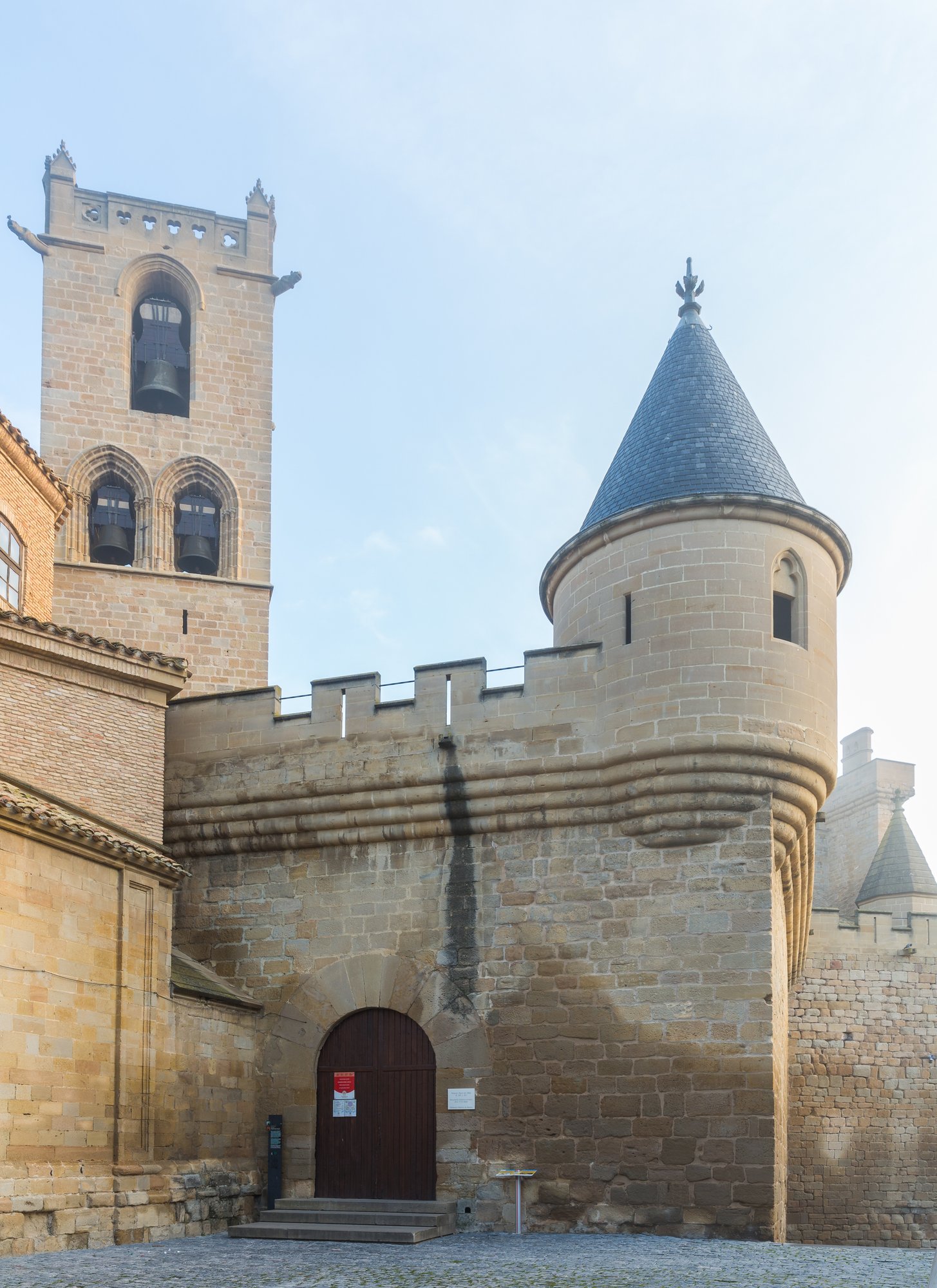A stone castle with a round slate-roofed tower and an arched doorway, a square bell tower rising behind it