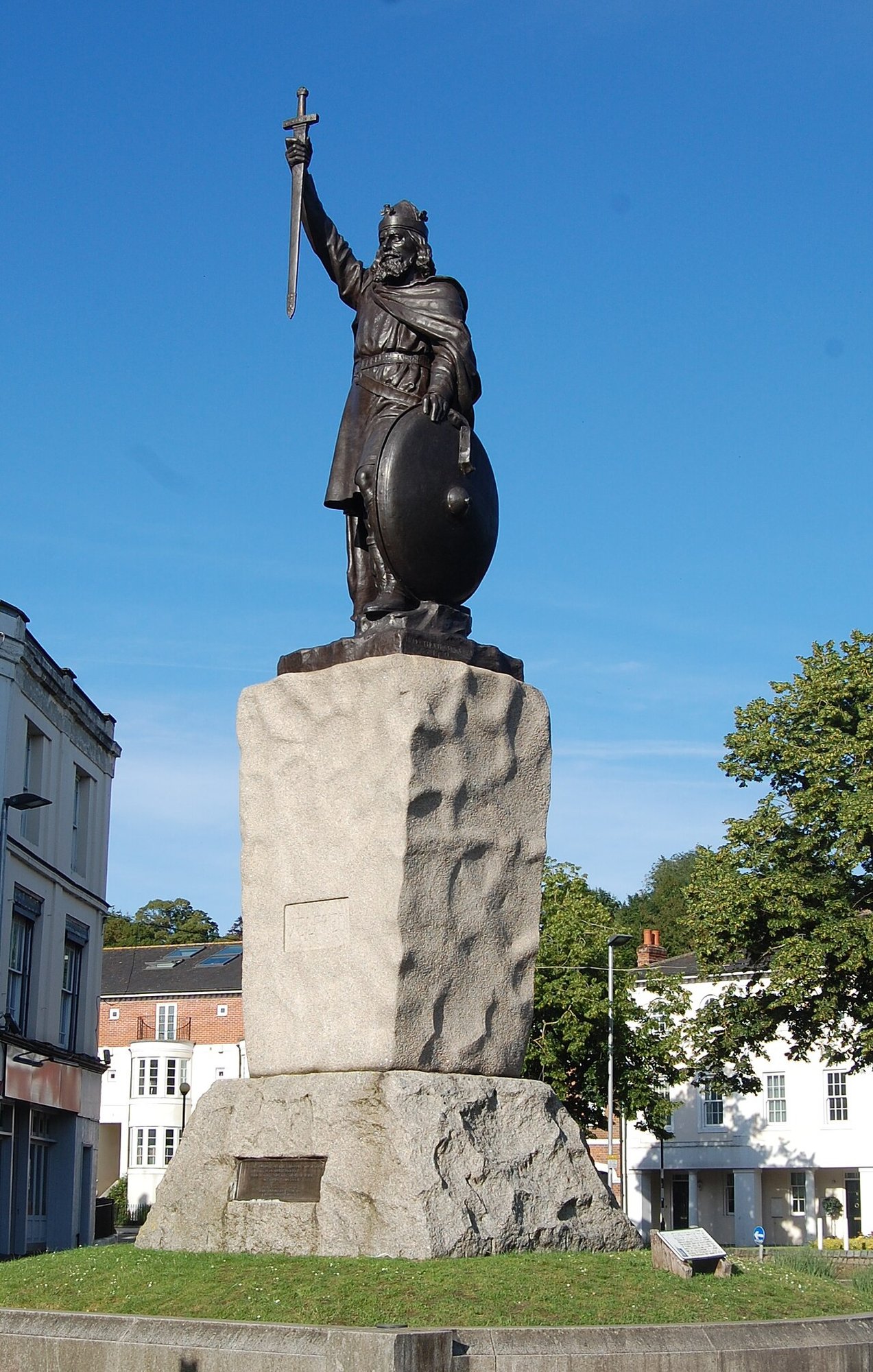 Bronze statue of a crowned king raising a sword, on a tall stone plinth against a blue sky with buildings behind