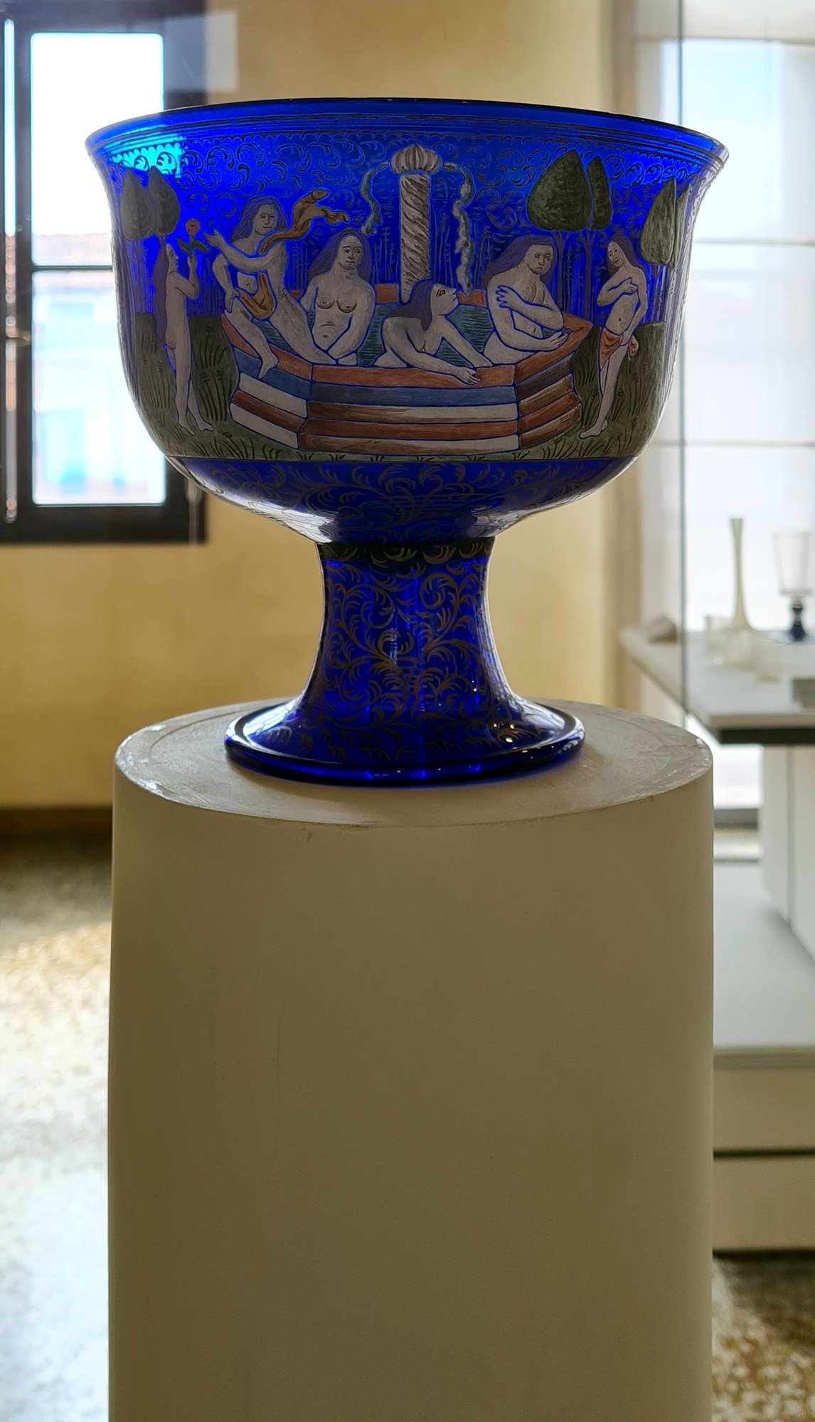 A deep blue glass footed bowl with white figural decoration, displayed on a plinth in a gallery