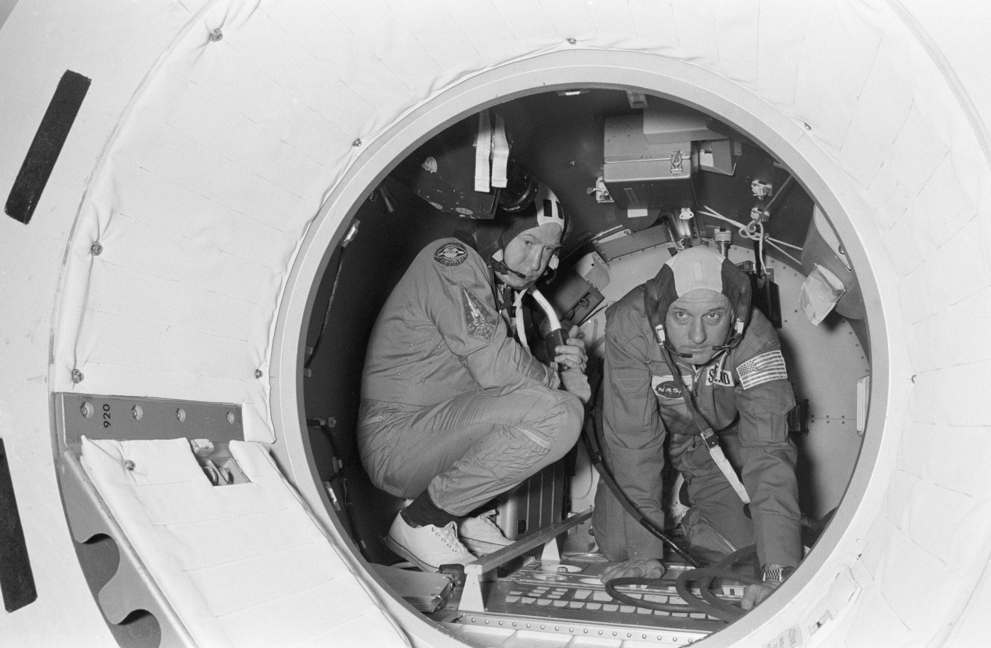 Two crew members in flight suits work inside the Apollo-Soyuz docking module, framed by its circular hatch