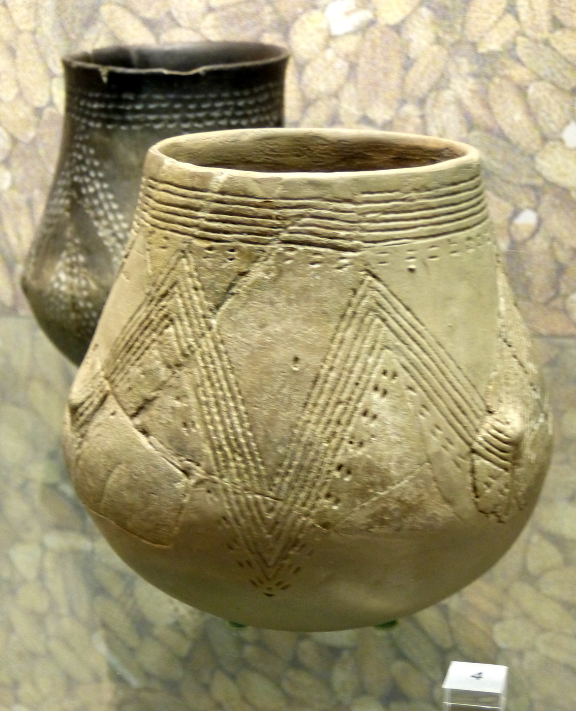 A round Linear Pottery culture vessel in a museum case, its body incised with bands of chevrons and rows of dots