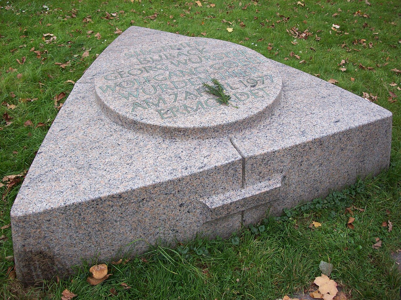 A granite memorial stone in grass, its inscription naming Siegfried Buback, Georg Wurster and Wolfgang Goebel, murdered on 7 April 1977