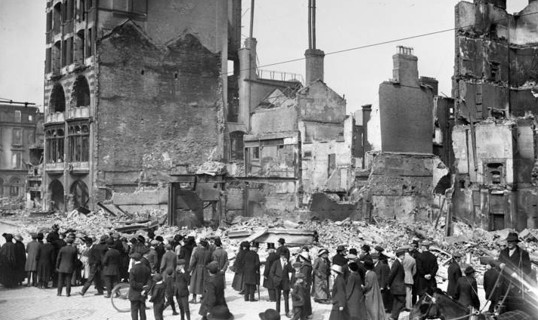 Historic photograph of a crowd standing among the gutted shells of buildings in a bombarded city street