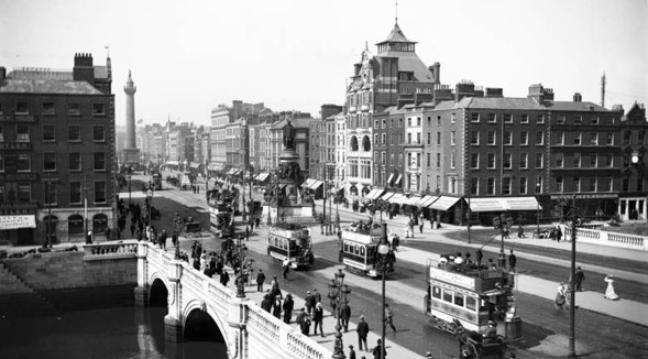 Historic photograph of a busy Dublin quayside street of tall buildings, with trams, awnings and crowds on the bridge